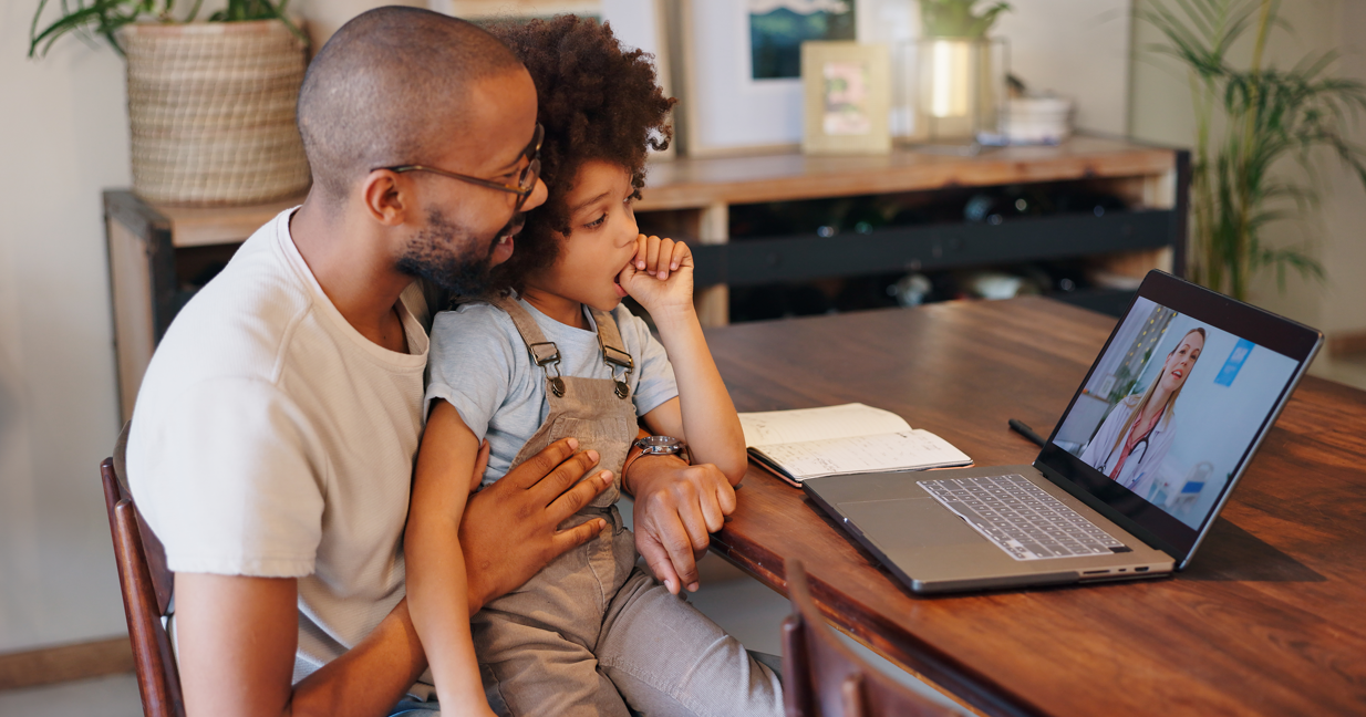 Dad with young son on his lapping talking to doctor via telehealth.